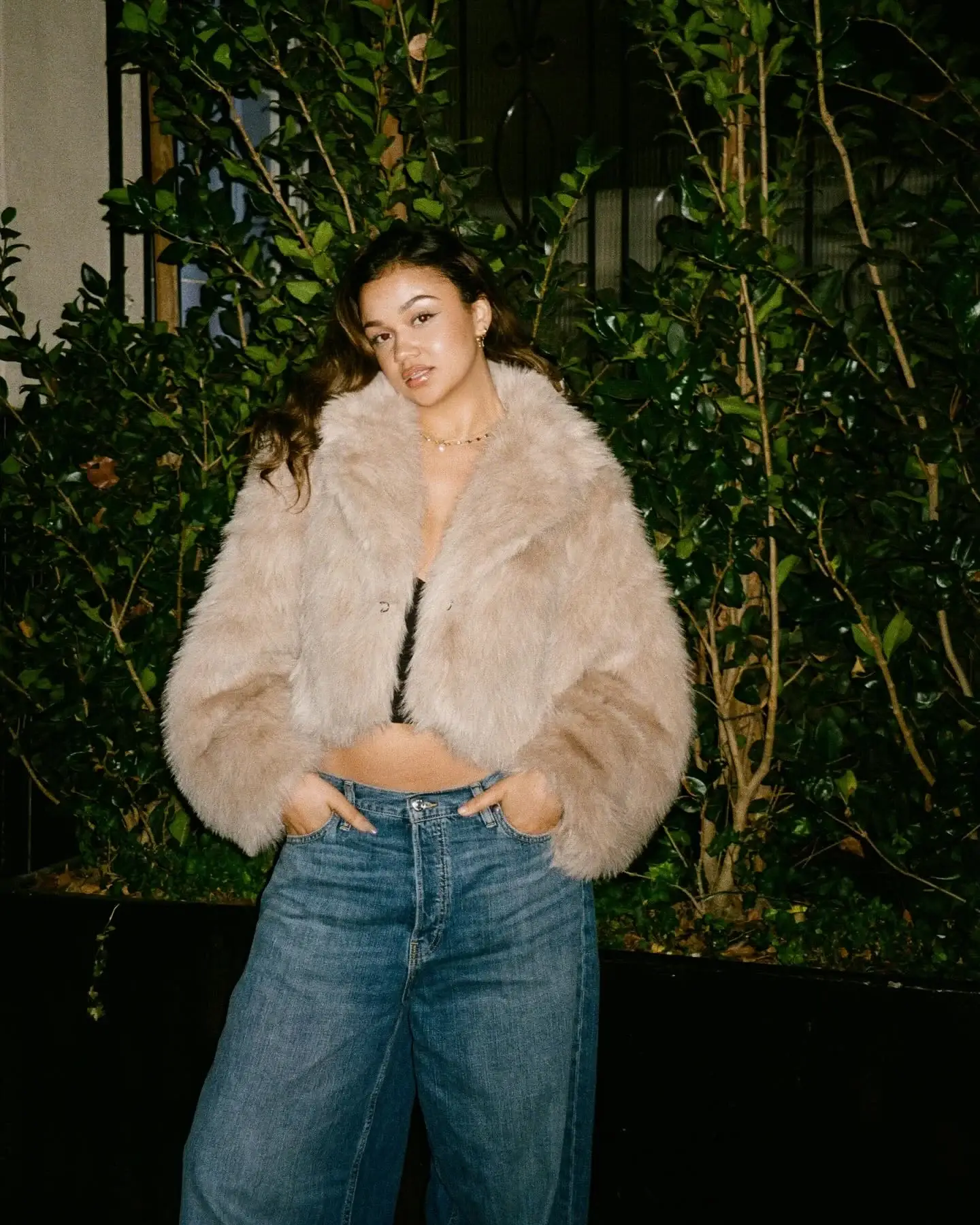 Madison Bailey poses outdoors at night wearing a beige cropped faux fur jacket and wide-leg denim jeans in front of green foliage.