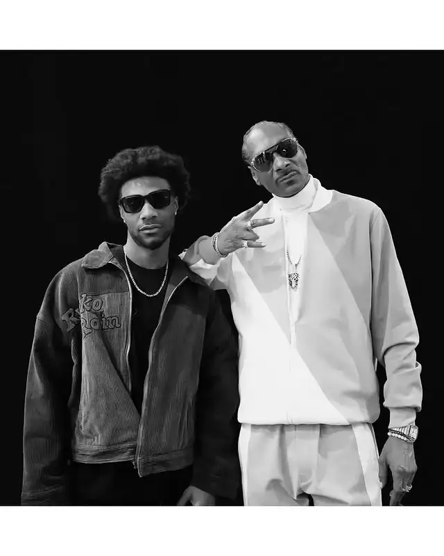 A black-and-white portrait of Jonathan Daviss and Snoop Dogg posing together in sunglasses against a dark background.