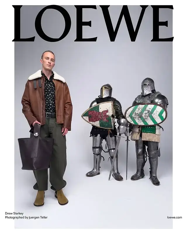 Drew Starkey posing in a shearling leather jacket with a handbag alongside two knight armors for a Loewe fashion campaign.
