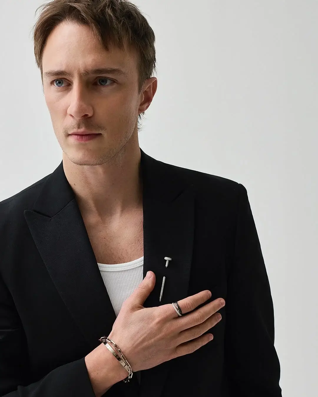 Drew Starkey posing in a black blazer and white tank top adorned with Cartier jewelry against a plain light background.