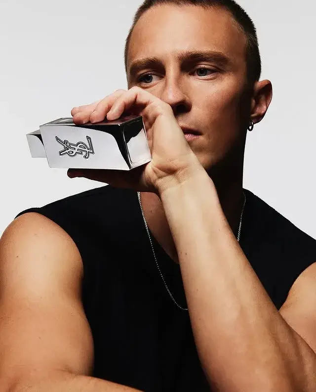 Drew Starkey posing in a sleeveless black shirt while holding a silver metallic Yves Saint Laurent fragrance bottle near his face.