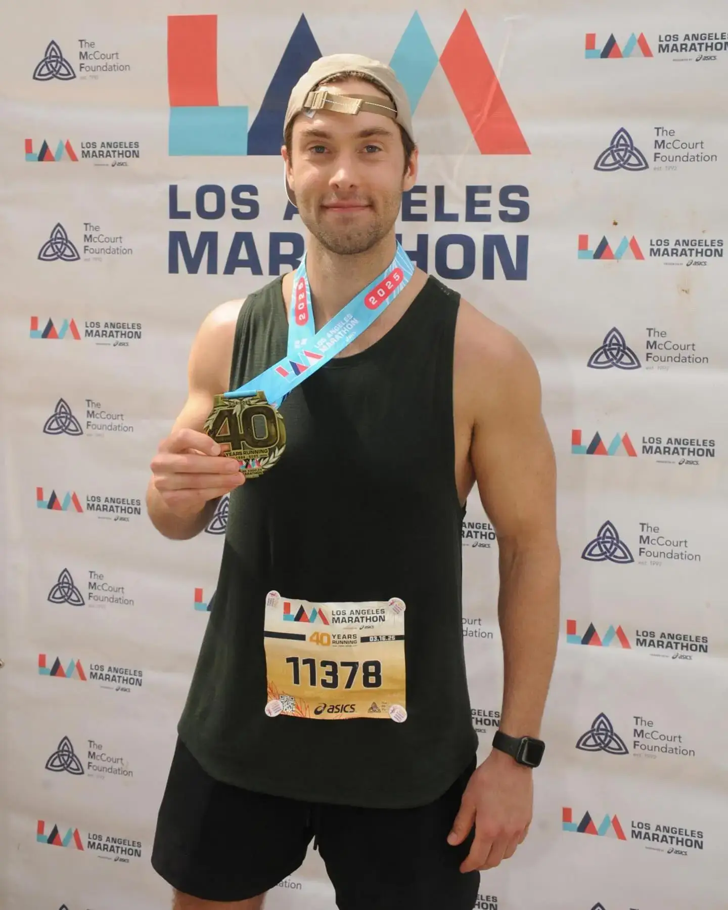 Austin North wearing a black tank top and bib number smiles proudly while holding up his finisher medal at the Los Angeles Marathon.
