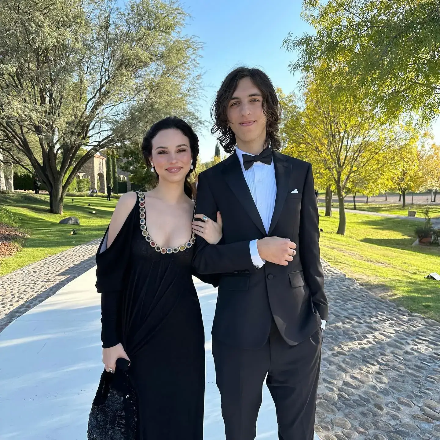 Fiona Palomo in an embellished black evening gown poses arm-in-arm outdoors with Luca Estrada wearing a classic black tuxedo and bow tie.