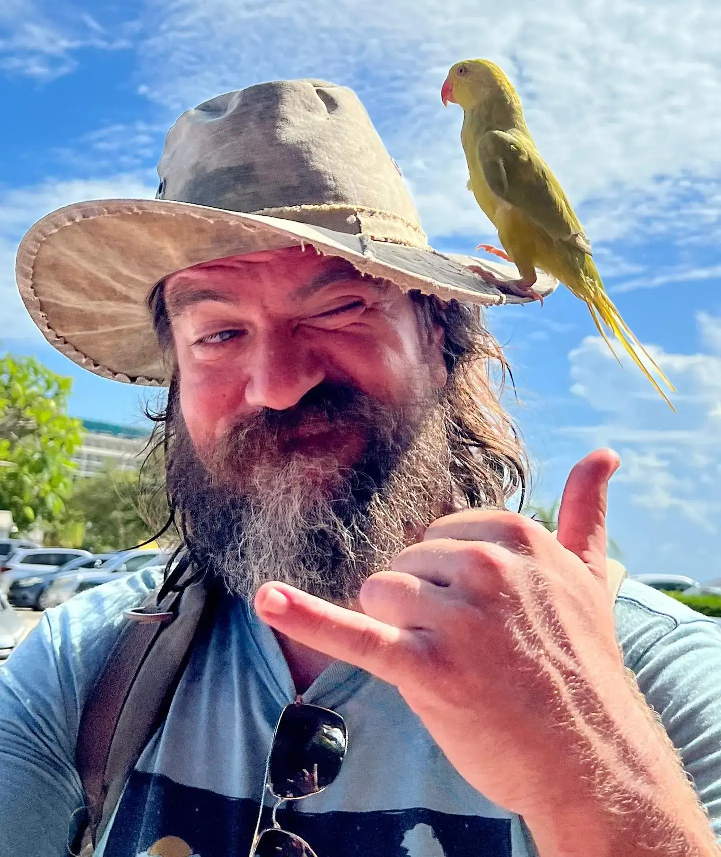 Charles Halford winking and giving a shaka hand sign with a small green parrot perched on his sun hat.