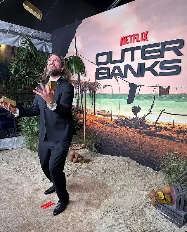 Charles Halford in a black suit jests by tossing gold bars into the air at a themed Netflix Outer Banks event.