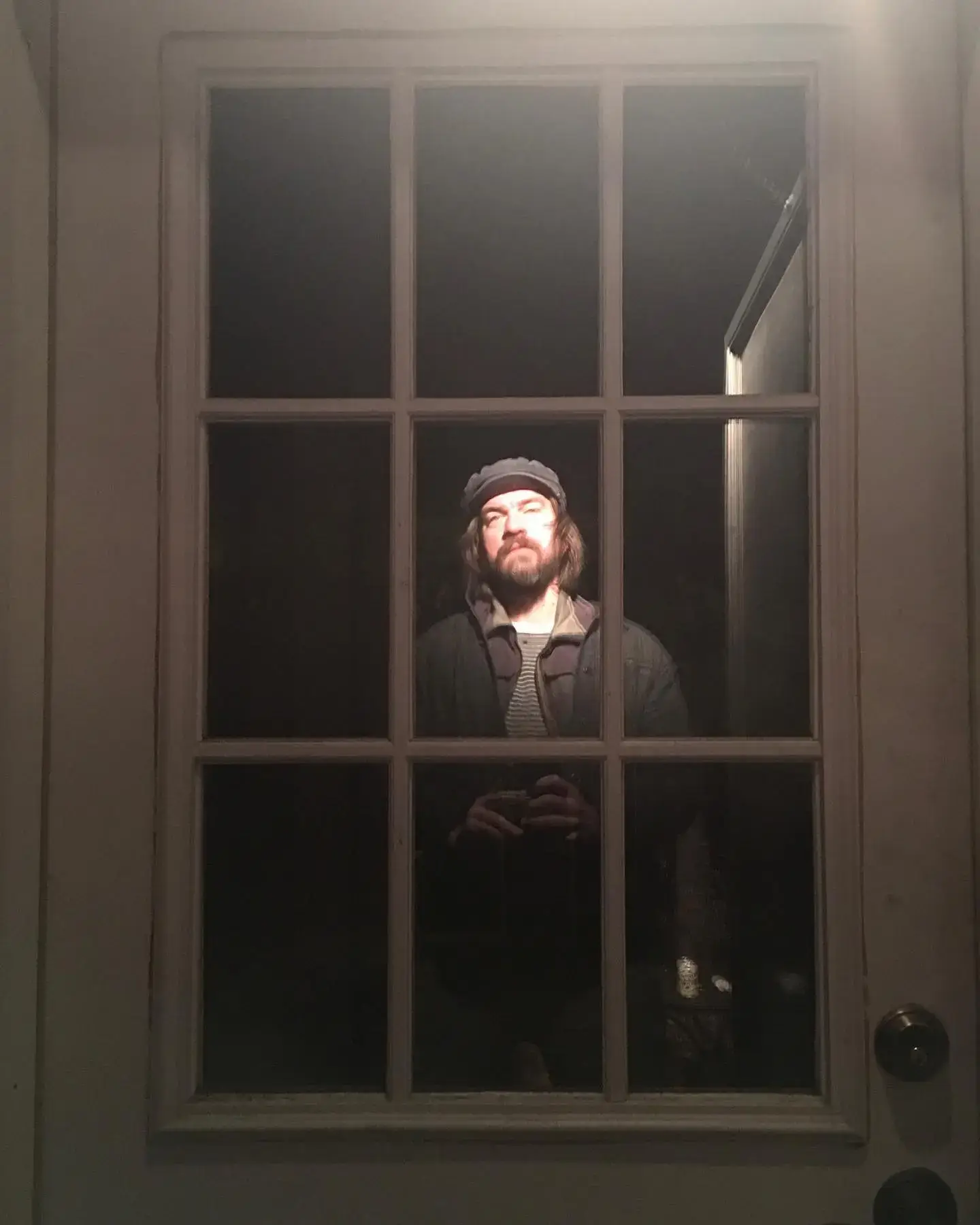 Charles Halford standing tall behind a multi-pane glass door wearing a cap and jacket in dramatic lighting.