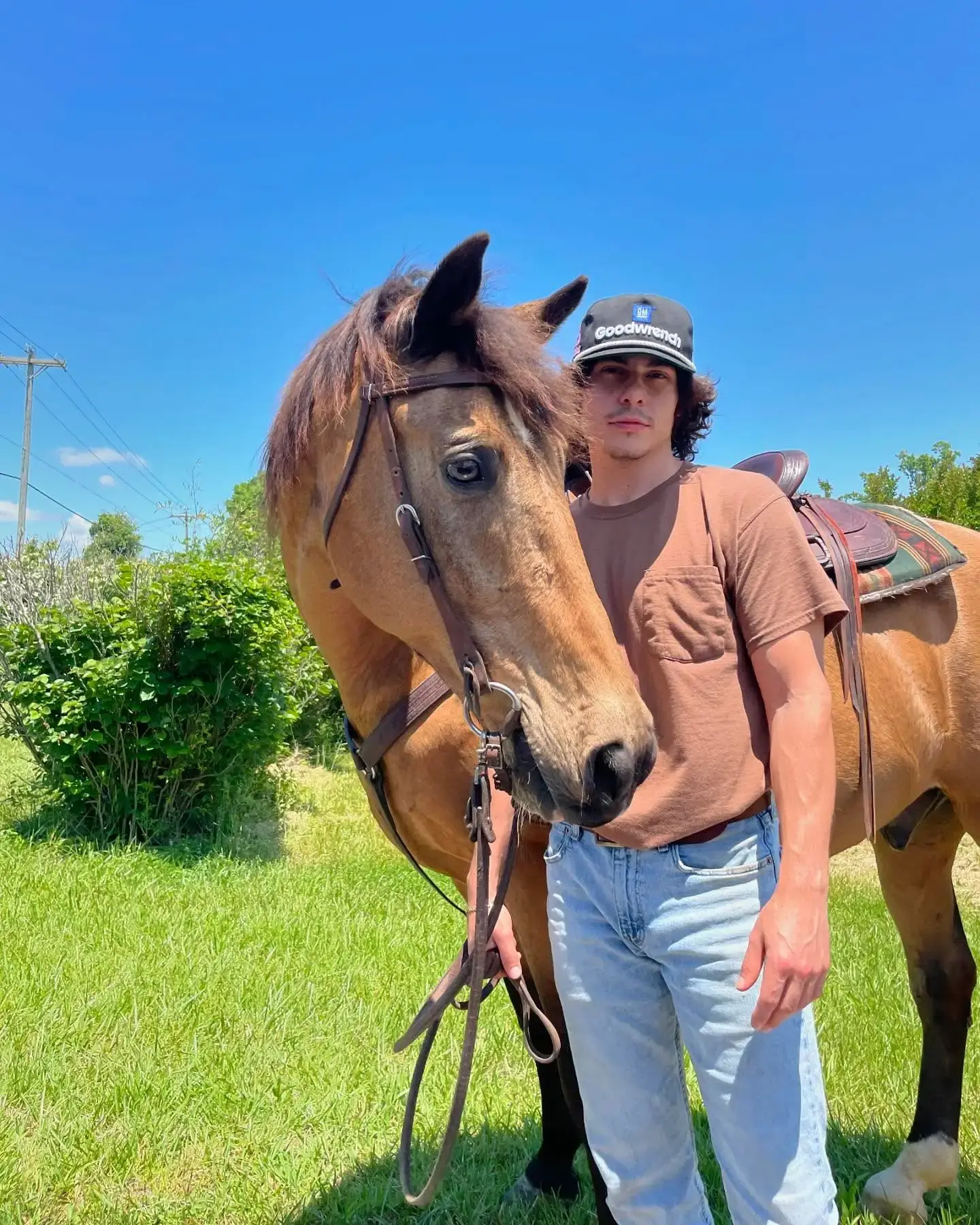 Nicholas Cirillo standing outdoors in a brown t-shirt and jeans next to a saddled horse in a sunny green field.
