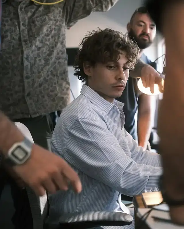 Nicholas Cirillo sitting in a striped dress shirt surrounded by crew members on a busy film production set.