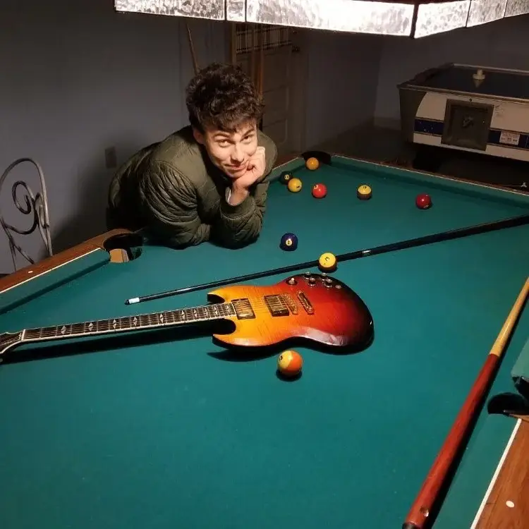 Nicholas Cirillo leaning over a green pool table featuring billiard balls, a cue stick, and an electric guitar.