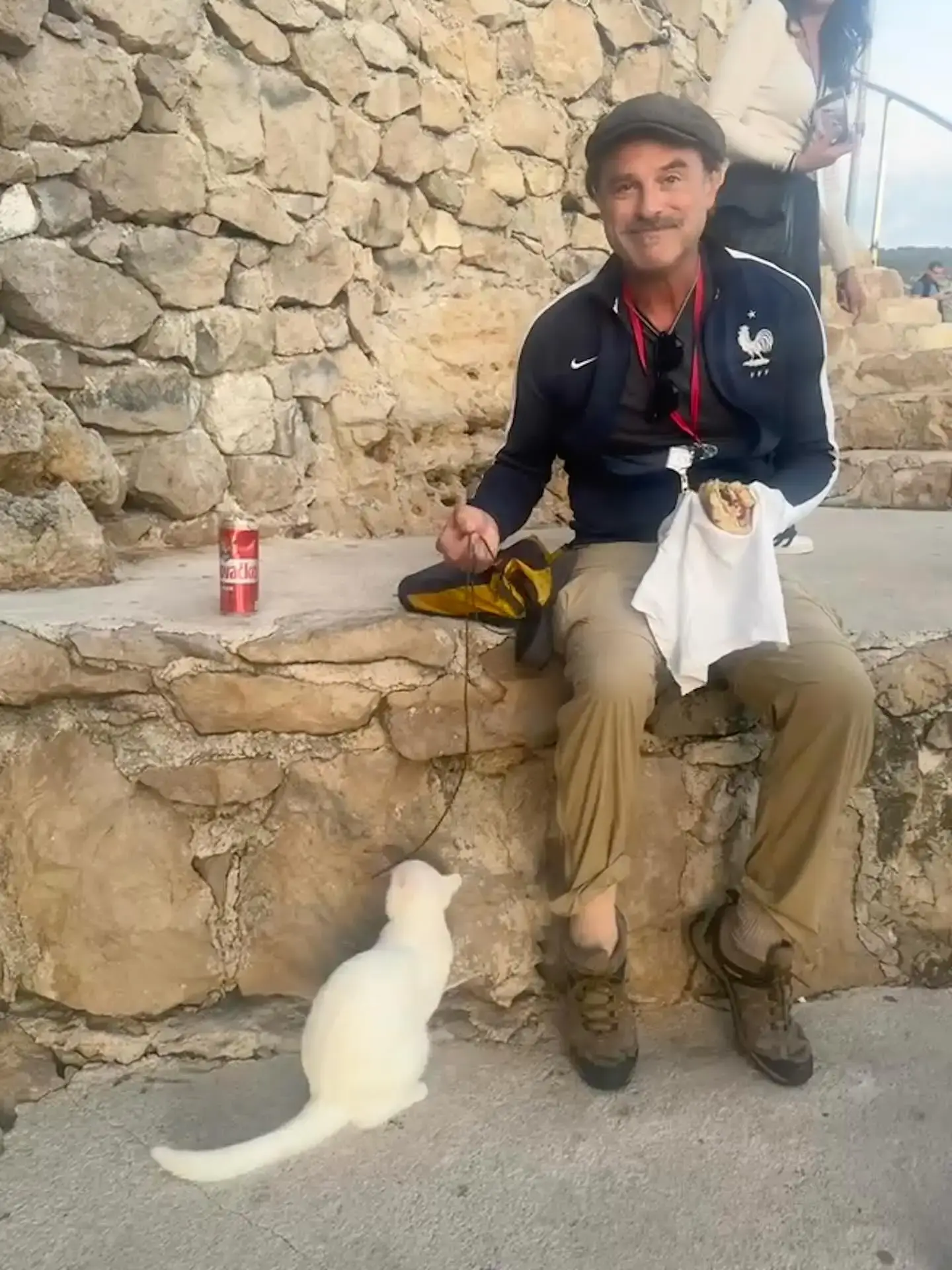 J. Anthony Crane sitting outdoors on a stone ledge in a cap and track jacket, smiling as he plays with a white cat.