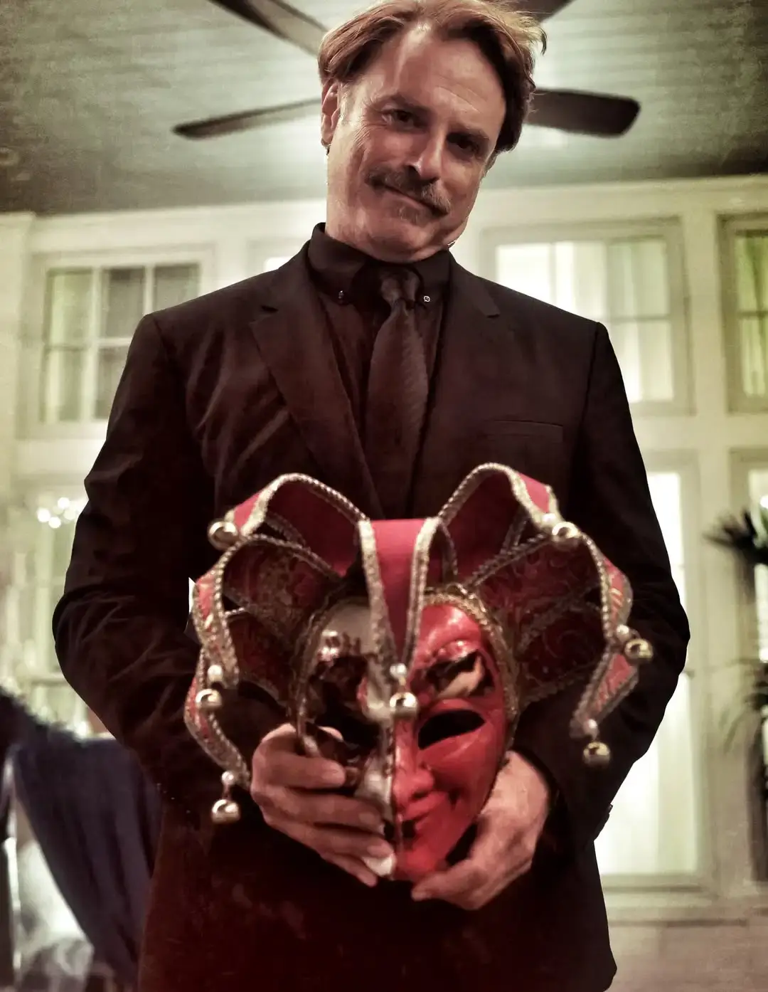 J. Anthony Crane dressed in a sharp dark suit holding an ornate red and gold Venetian jester mask.