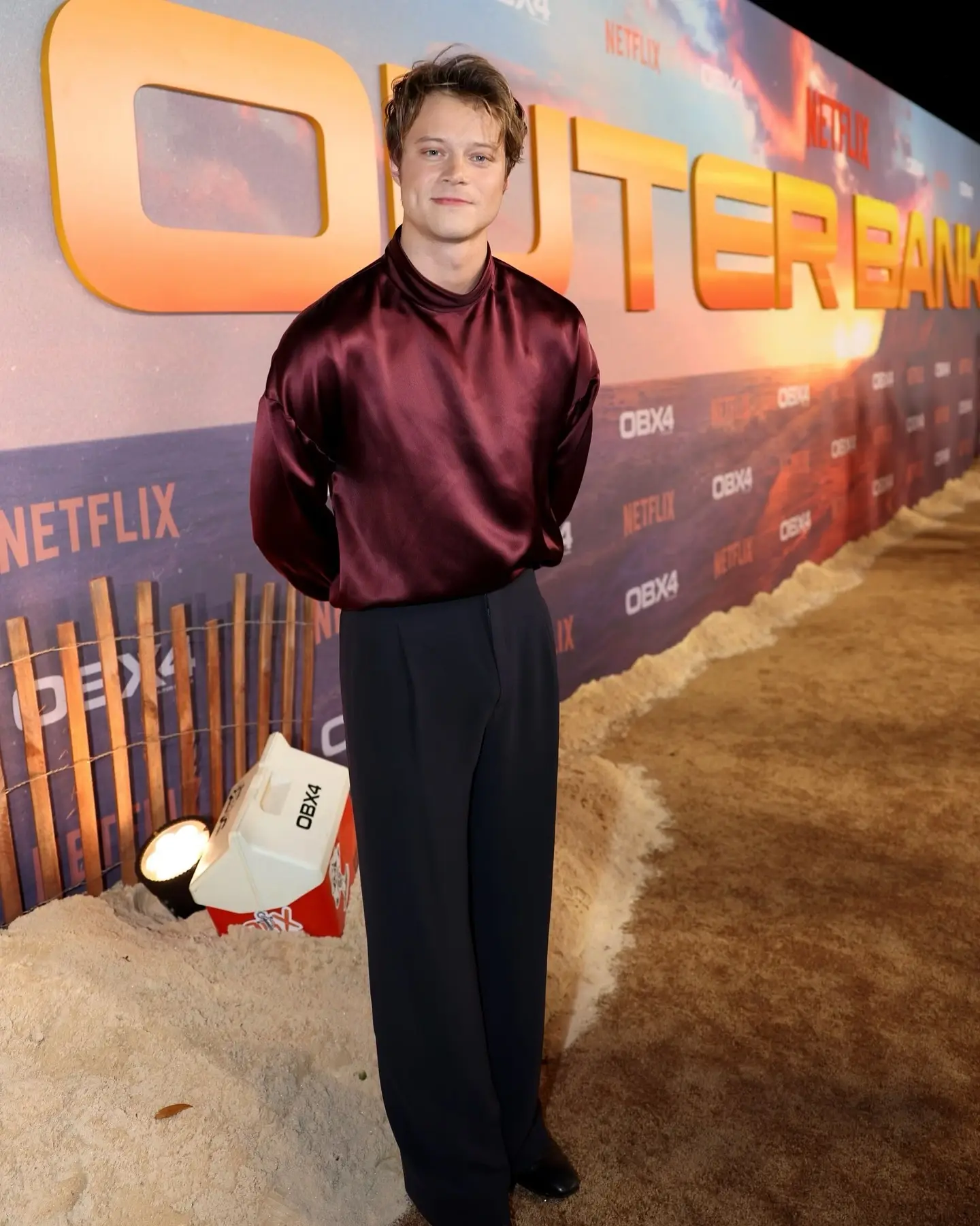 Rudy Pankow posing full-length in a maroon satin high-neck blouse and wide-leg black trousers on the Outer Banks red carpet.
