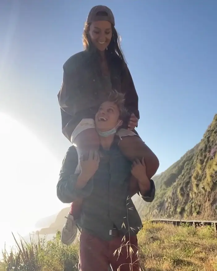 Rudy Pankow happily carrying his girlfriend Elaine Siemek on his shoulders outdoors along a scenic cliffside road.