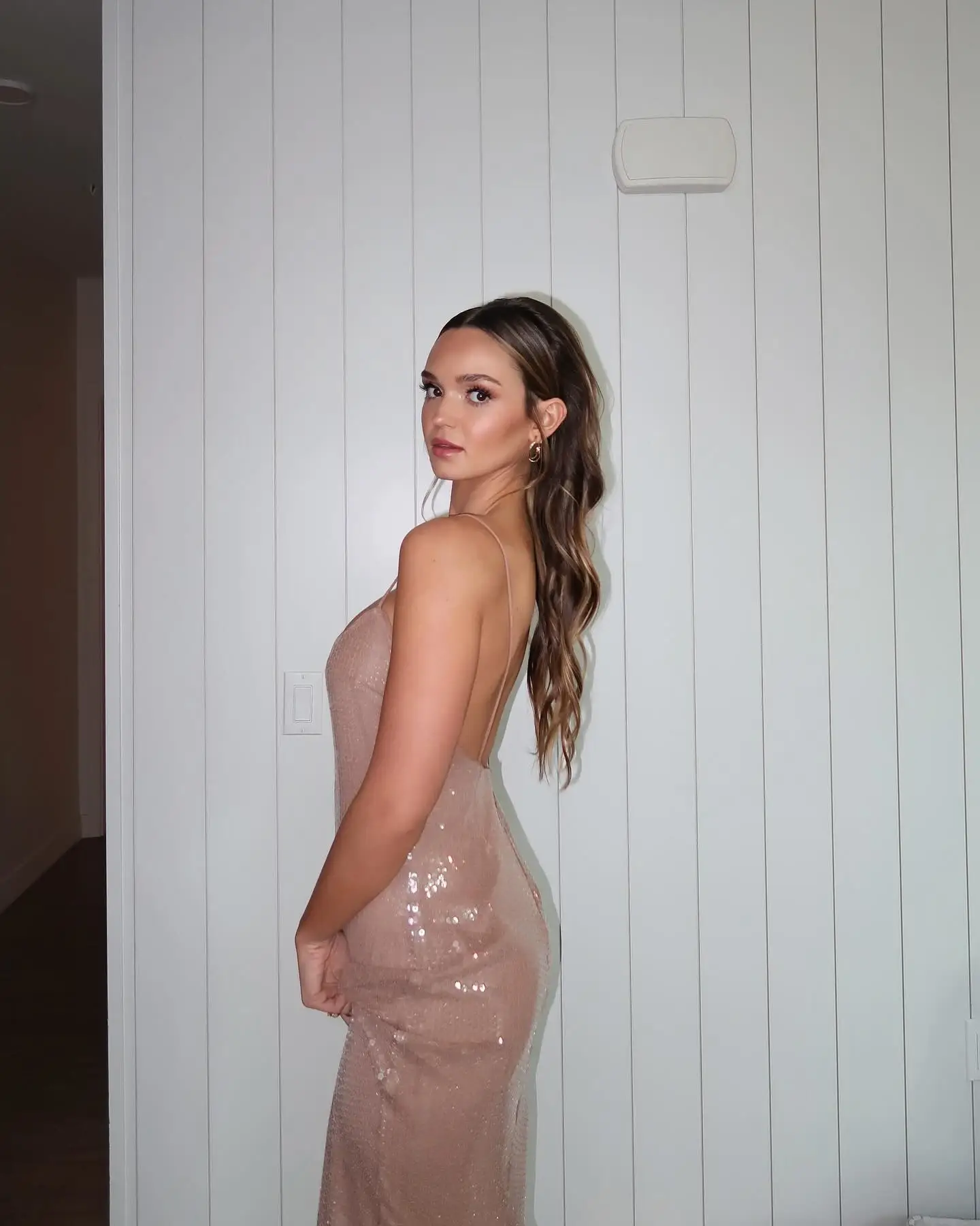 Lilah Pate posing in a side profile wearing a sequined champagne-pink open-back dress against a white paneled wall.