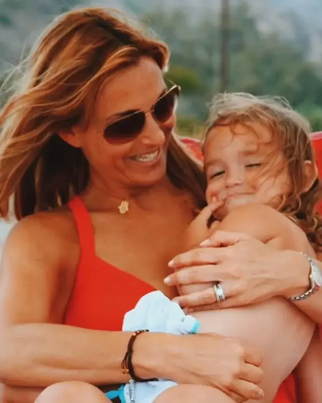 Lilah Pate as a young toddler smiling lovingly while being held by her mother, Jennifer Pate, in sunglasses and a red swimsuit.