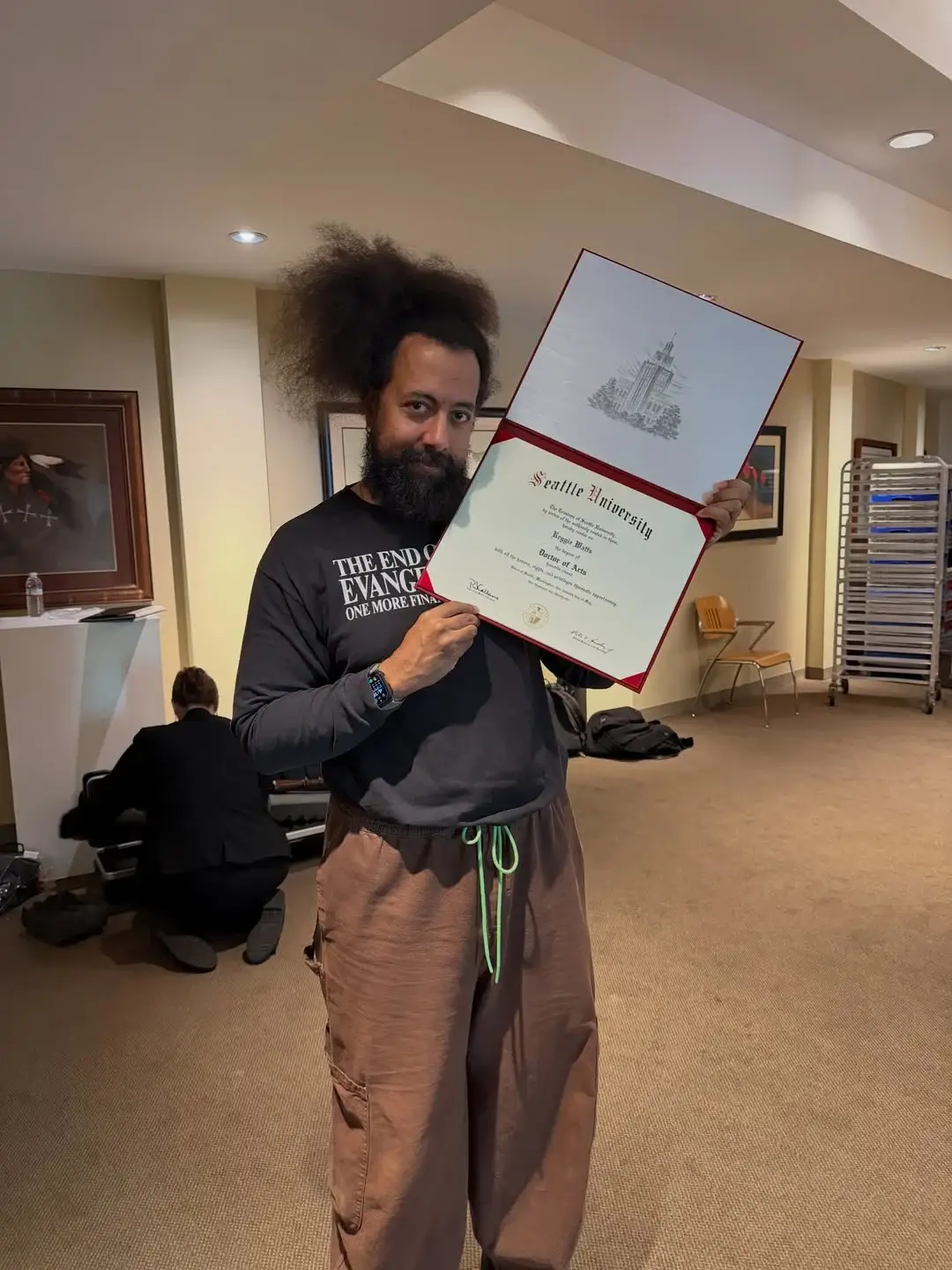 Reggie Watts proudly holding up an open honorary doctorate diploma folder from Seattle University indoors.