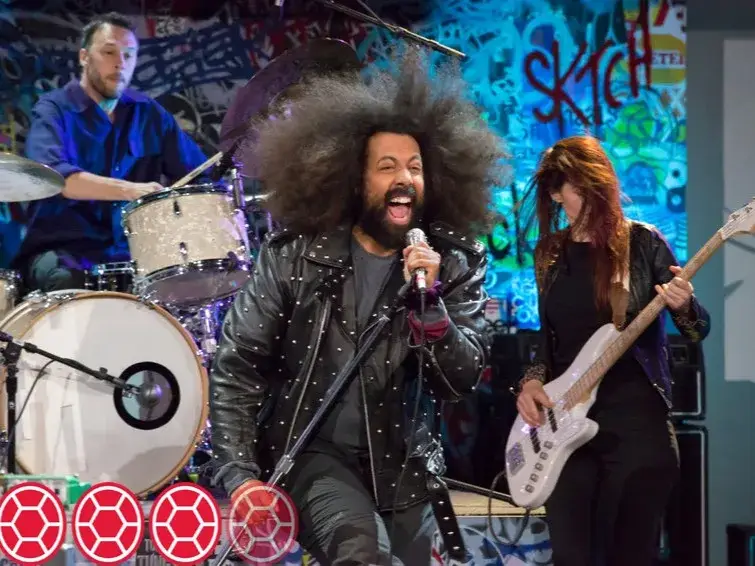 Reggie Watts passionately singing into a microphone in a studded black leather jacket with a live band performing on stage.