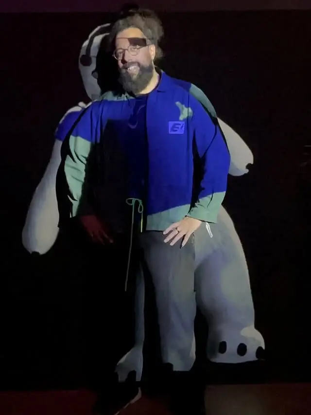 Reggie Watts smiling in glasses and a blue and green jacket with an animated character projection cast over him.
