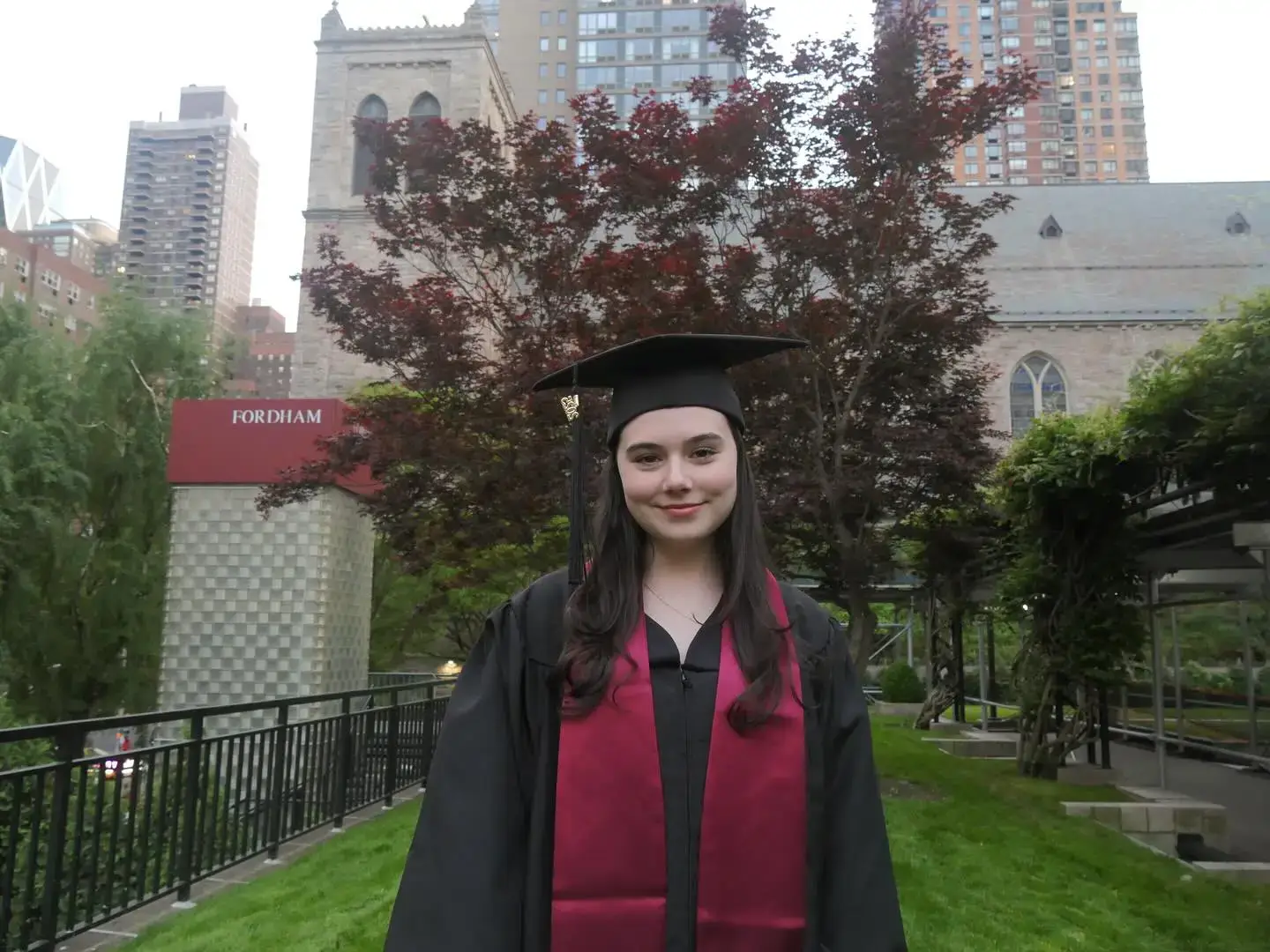 Julia Antonelli smiling outdoors in a black cap and graduation gown with a maroon stole at Fordham University.