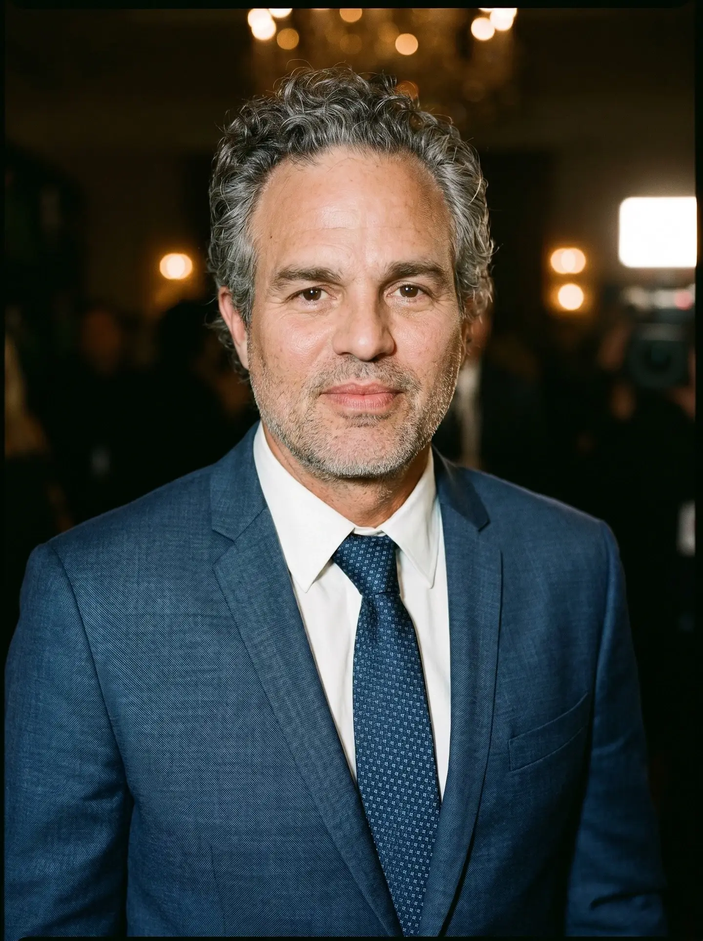 Mark Ruffalo posing in a tailored blue suit and patterned tie against a softly lit evening background.