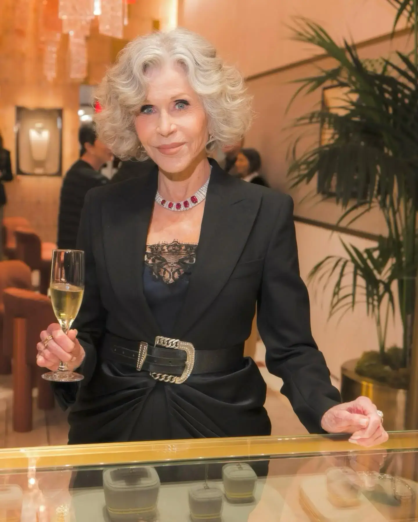 Jane Fonda holding a glass of champagne while posing in a tailored black suit with an ornate statement necklace and belt.