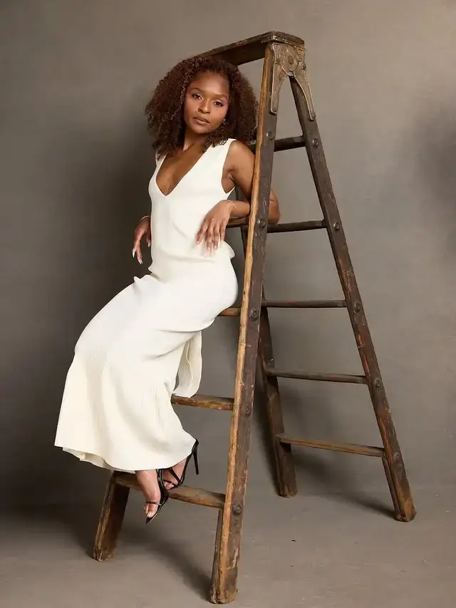Dominique Thorne wearing an ivory sleeveless maxi dress while seated gracefully on a rustic wooden ladder against a grey studio backdrop.