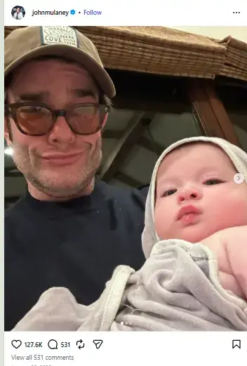 John Mulaney smiling playfully in a cap and glasses while holding his baby daughter Méi June Mulaney wrapped in a hooded towel.