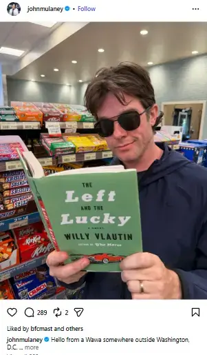 John Mulaney wearing dark sunglasses while holding and reading a copy of Willy Vlautin