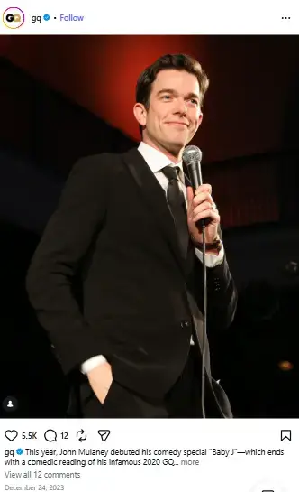 John Mulaney smiling on stage in a sharp black suit and tie while holding a microphone.
