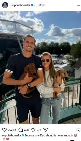 Alexander Zverev and his gf Sophia Thomalla standing on a sunny balcony, each holding a brown dachshund puppy.