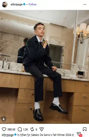 Elliot Page posing in a sleek dark jacket, trousers, white socks, and loafers while seated on a marble bathroom vanity.