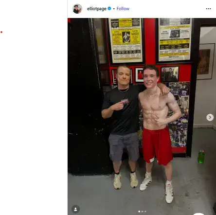 A smiling, shirtless Elliot Page in red boxing shorts posing beside his trainer inside a gym.