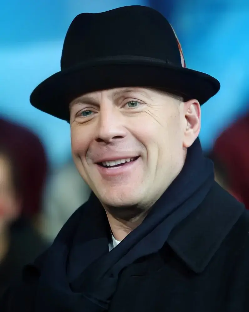 Close-up portrait of Bruce Willis smiling while wearing a black fedora hat, scarf, and dark coat.