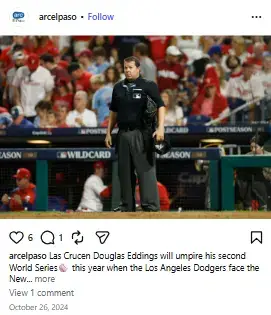 MLB umpire Doug Eddings standing on the baseball field in full uniform with crowded stadium stands behind him.