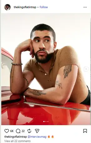 Bad Bunny leaning his arm on a red car while resting his chin on his hand in a brown knit polo shirt.