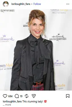 Lori Loughlin smiling in a black blazer and pussy-bow blouse against a Hallmark Channel red carpet backdrop.