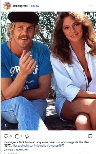 Young Nick Nolte wearing a cap and holding a cigarette while sitting outdoors beside Jacqueline Bisset on the set of The Deep.