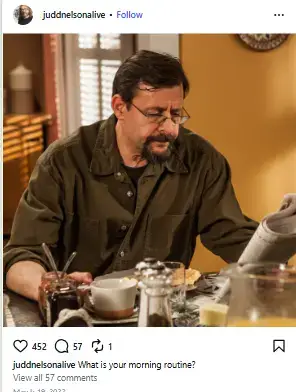 Judd Nelson wearing eyeglasses and an olive green button-up shirt while reading a newspaper at a breakfast table.