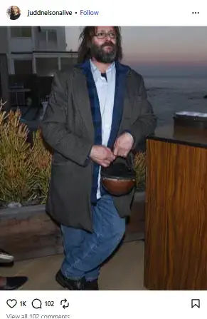 Full-length photo of Judd Nelson sporting a full beard, long coat, and jeans while standing outdoors near a coastal patio.