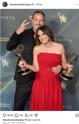 Mariska Hargitay holding two Emmy awards in a strapless red gown while her husband Peter Hermann smiles and makes a peace sign behind her.