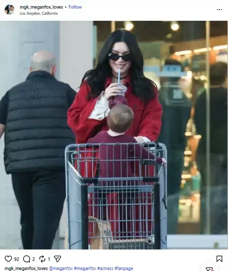 Megan Fox smiling in sunglasses and a red coat while pushing her newborn daughter in a shopping cart