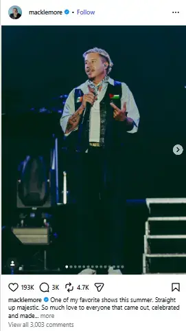 Macklemore performing live onstage holding a microphone while wearing a patched vest and neck scarf.