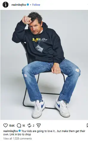 Michael J. Fox sitting on a black chair wearing a dark hoodie, blue jeans, and sneakers against a white background.