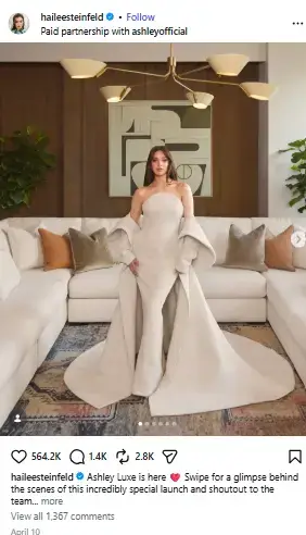 Hailee Steinfeld standing in an elegant off-white strapless gown with a dramatic cape inside a stylish modern living room.