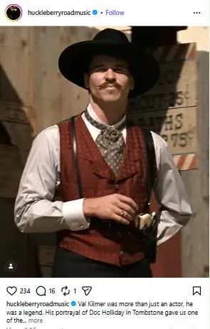 Val Kilmer as Doc Holliday standing in a red vest and black cowboy hat in a scene from the film Tombstone.