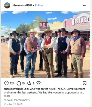 A group of historical reenactors in Old West cowboy attire posing outdoors alongside a news reporter in front of the O.K. Corral sign.