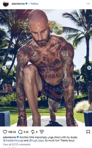 Shirtless Adam Levine in a low lunge yoga pose on an outdoor mat, showing extensive tattoos covering his chest, arms, and stomach.