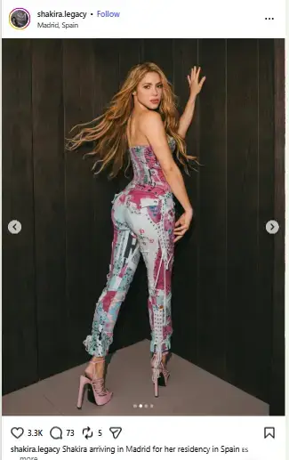 Shakira posing against a dark wooden wall wearing a patterned pink and light blue strapless jumpsuit with high-heeled platform sandals.