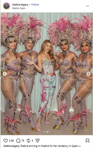 Shakira in a floral print bustier and trousers smiling in the center of four carnival dancers wearing elaborate pink feathered headdresses.