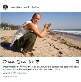 Woody Harrelson crouching barefoot on a sandy beach with his hands pressed together near the ocean shoreline.